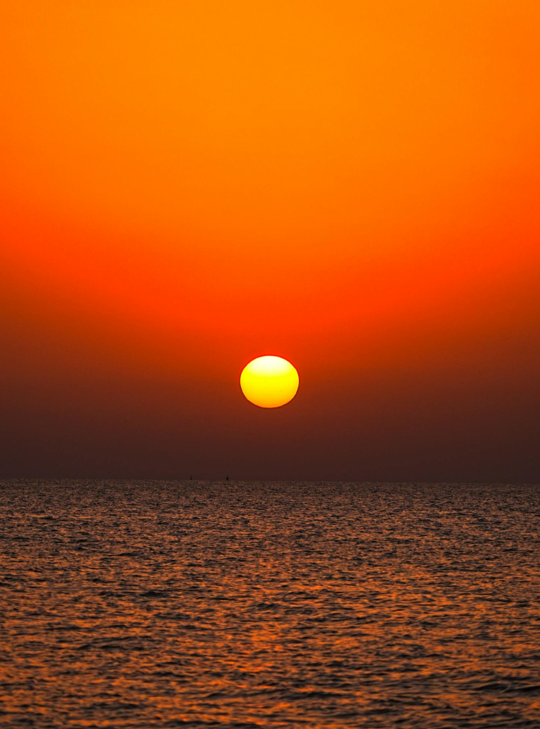 Glowing orange sunset over calm ocean horizon with golden sun reflecting on tranquil sea water.
