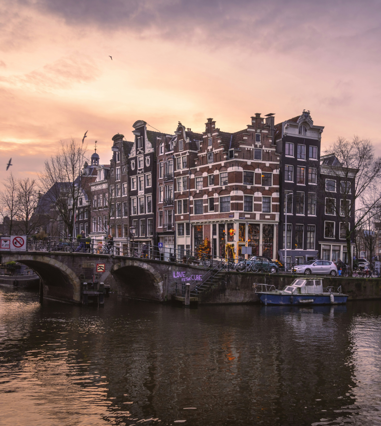 Historic canal houses and arched bridge in Amsterdam at sunset with boats and reflections on the water.