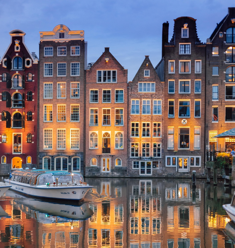 Traditional Amsterdam canal houses illuminated at dusk with their reflections in the water and a sightseeing boat passing by.