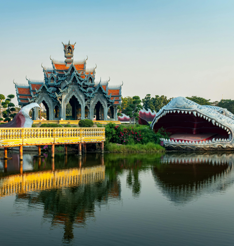 Ornate Thai pavilion at Ancient City in Samut Prakan with a large dragon-mouth sculpture beside a peaceful lake reflecting the temple architecture.