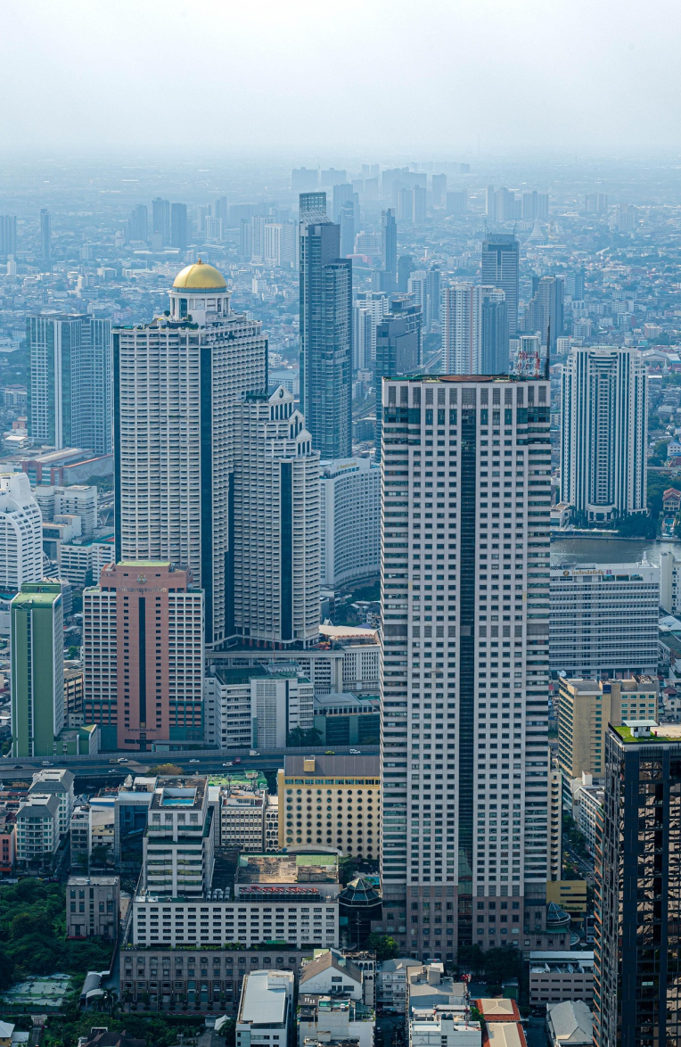 Aerial view of Bangkok’s skyline featuring tall modern high-rise buildings and dense urban architecture on a hazy day.