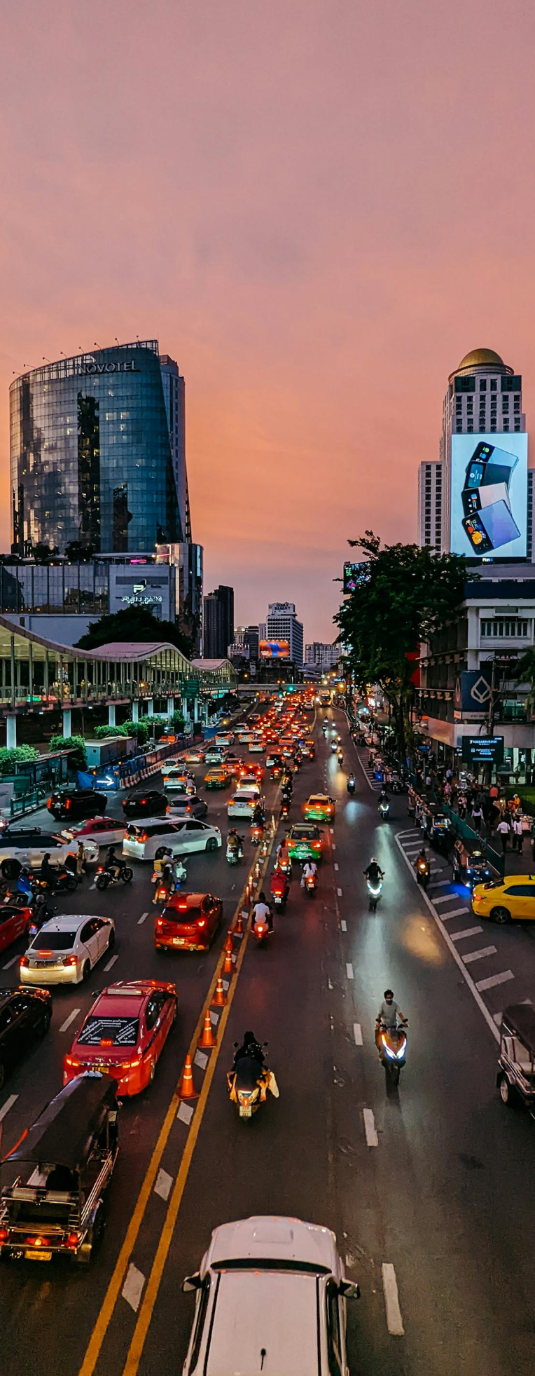 Busy Bangkok street at sunset with heavy traffic, motorbikes, tall modern buildings, and illuminated billboards along the Sukhumvit city skyline.