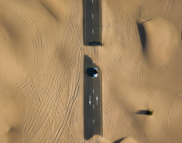 Aerial view of a car driving on a straight road through Dubai desert dunes with tire tracks.