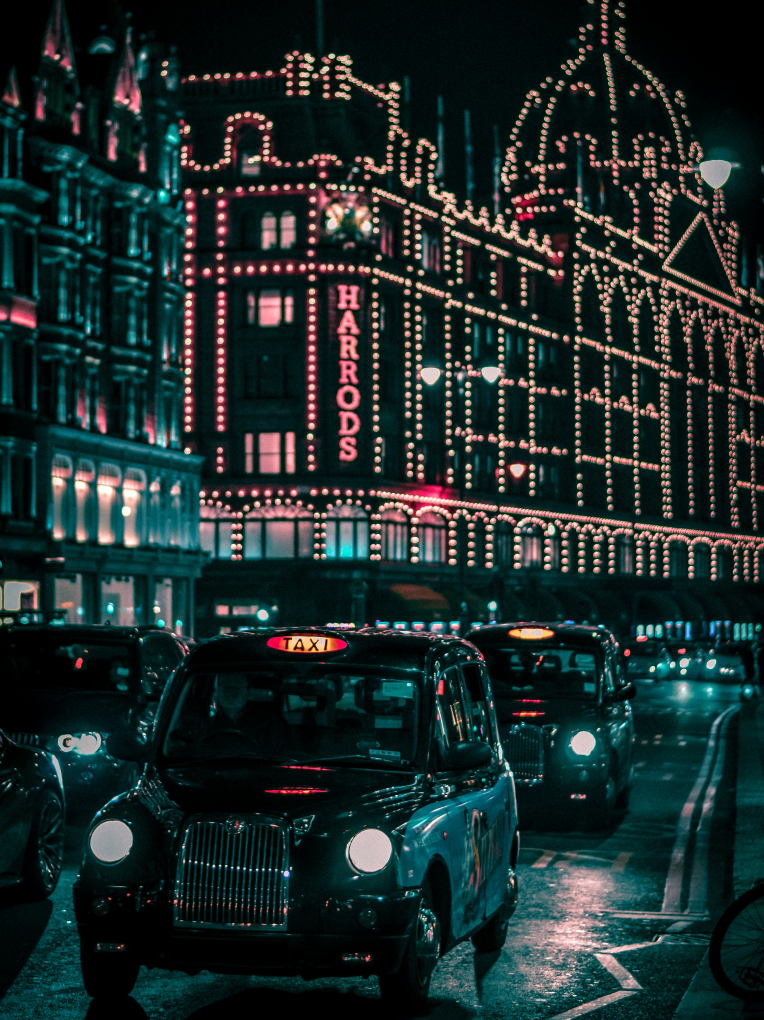 Harrods department store illuminated at night in Knightsbridge with classic London black cabs in traffic.