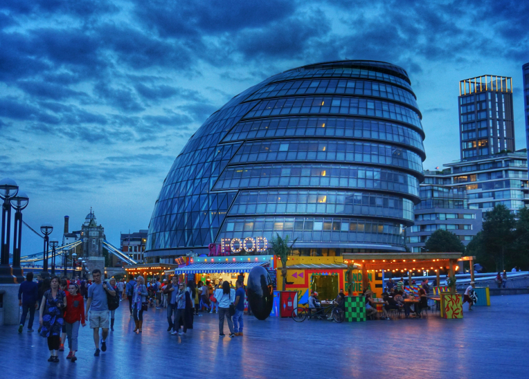 London City Hall on the South Bank at dusk, with lively food stalls and pedestrians, Tower Bridge visible by the River Thames.