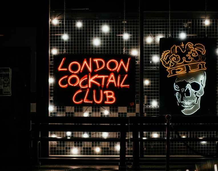 Neon sign reading “London Cocktail Club” with skull and crown artwork on a bar window at night.