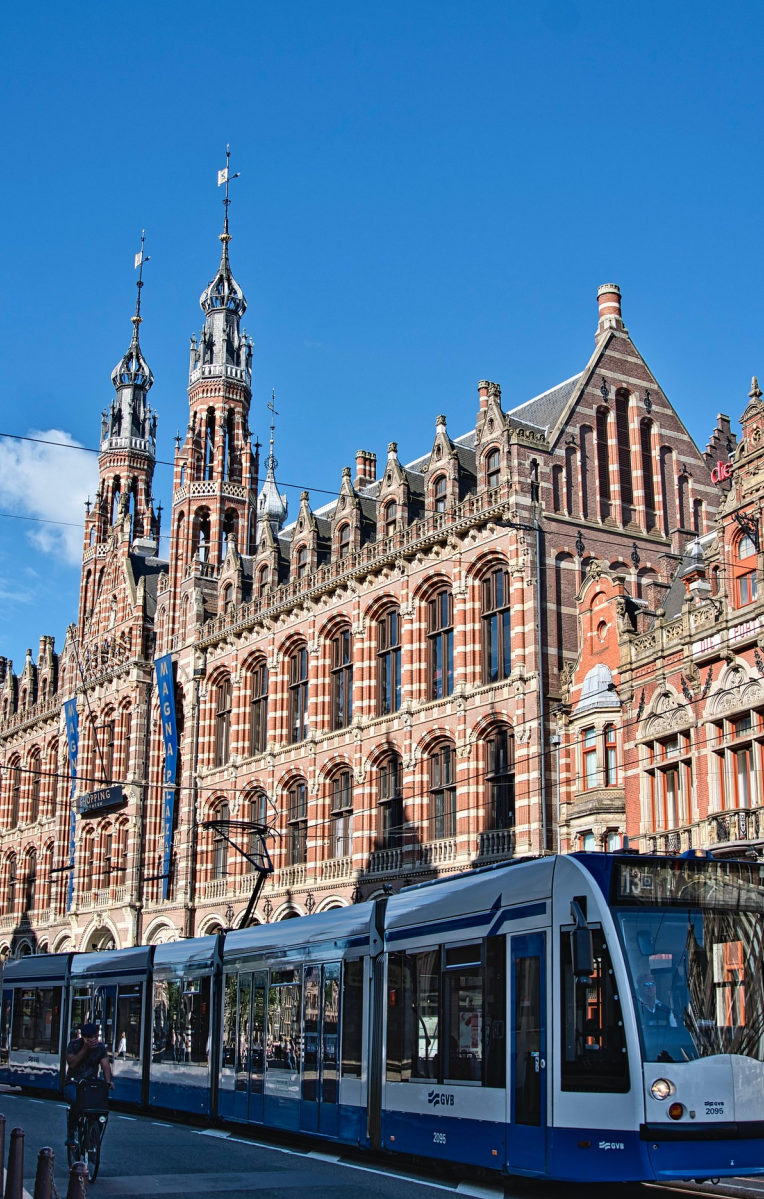 Historic Magna Plaza building in Amsterdam with a modern blue and white tram passing in front on a sunny day.