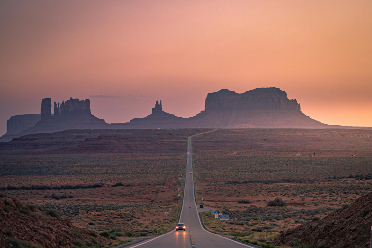 US-163 straight road leading to Monument Valley mesas at sunset in the Utah–Arizona desert.