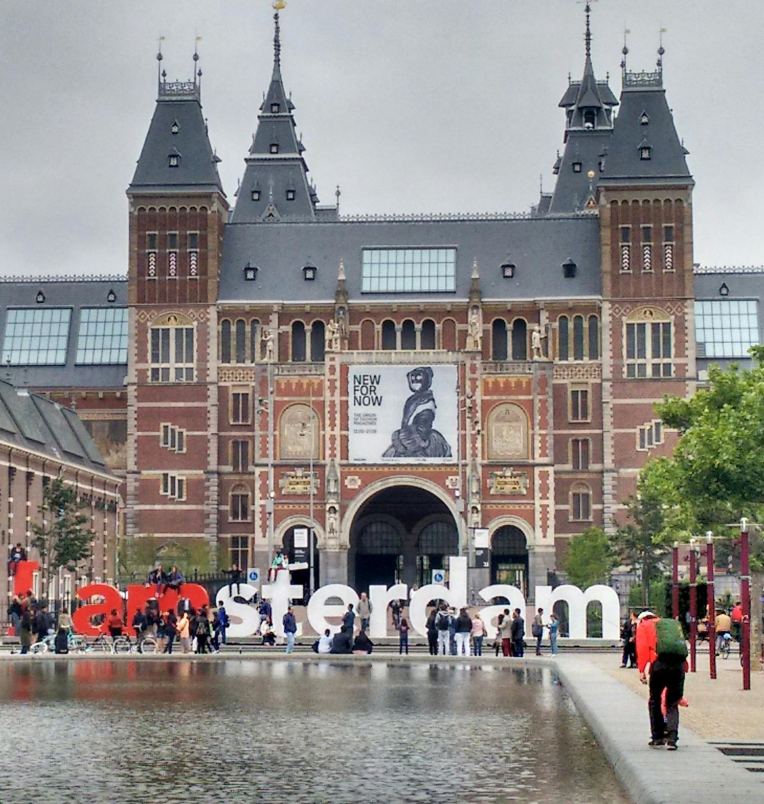 The famous I amsterdam sign in front of the Rijksmuseum with crowds and its reflection in the water on a cloudy day.