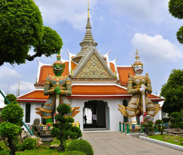 Two colorful guardian statues standing at the entrance of a Thai temple with an ornate roof and decorative details at Wat Arun in Bangkok.
