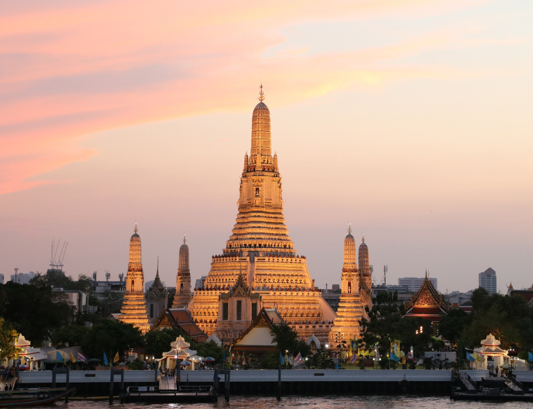 Wat Arun temple illuminated at sunset along Bangkok’s Chao Phraya River, showcasing its iconic central prang and surrounding spires.