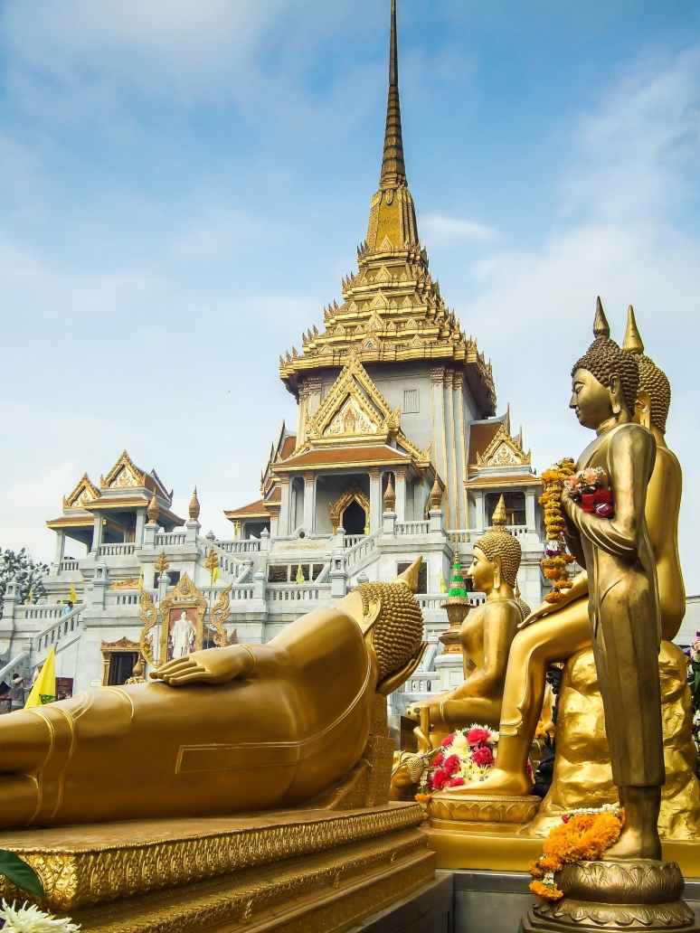 Golden Buddha statues in front of Wat Traimit’s ornate temple architecture in Bangkok, featuring traditional Thai design and a towering golden spire.
