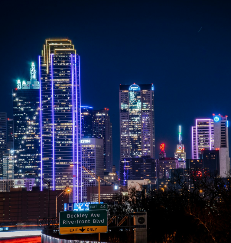 Night view of the downtown Dallas skyline featuring brightly lit skyscrapers, neon building outlines, and a highway exit sign in the foreground.