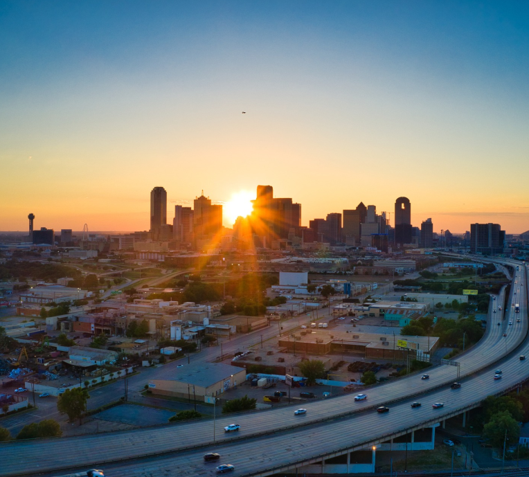 Aerial view of the Dallas, Texas skyline at sunrise with the sun rising behind downtown buildings and highways in the foreground.