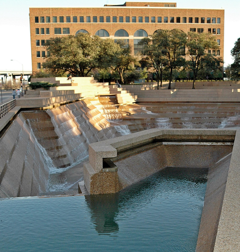 View of the Fort Worth Water Gardens cascade pool in downtown Fort Worth, Texas, featuring terraced concrete steps, flowing water, and surrounding trees.