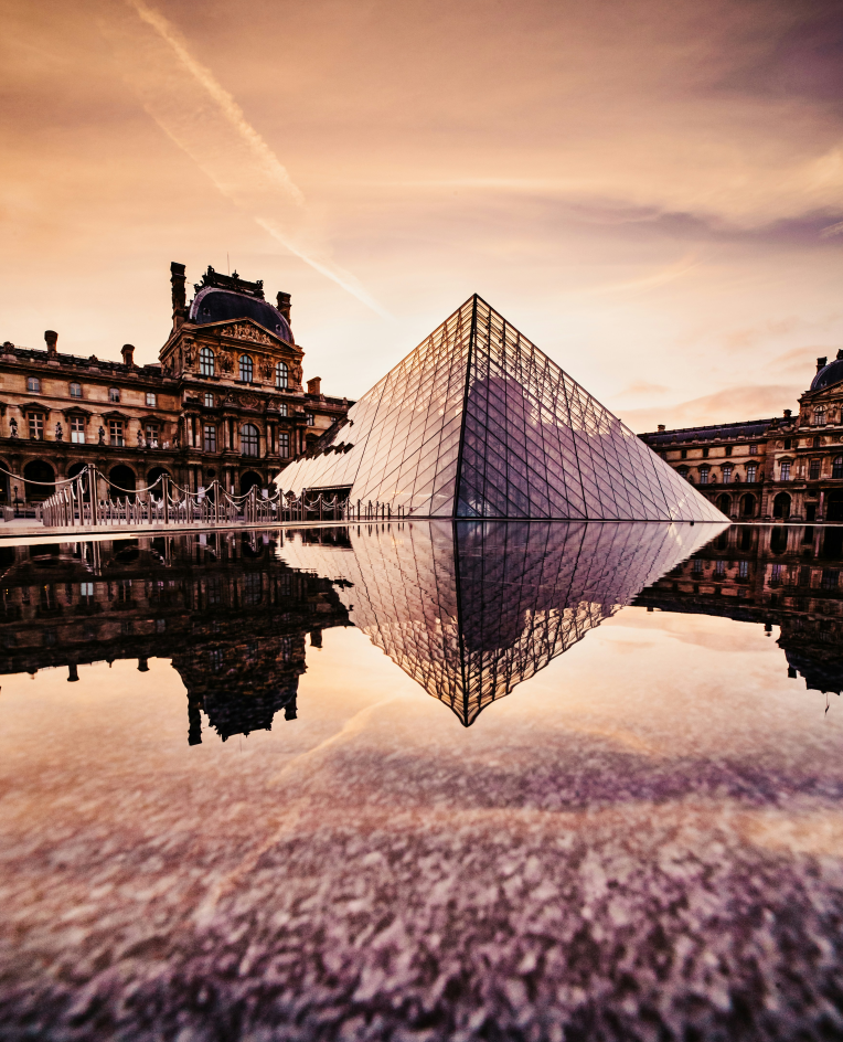 The Louvre Pyramid in Paris reflected in water at sunset, highlighting iconic attractions easily reached when staying near Paris-Charles de Gaulle Airport (CDG).