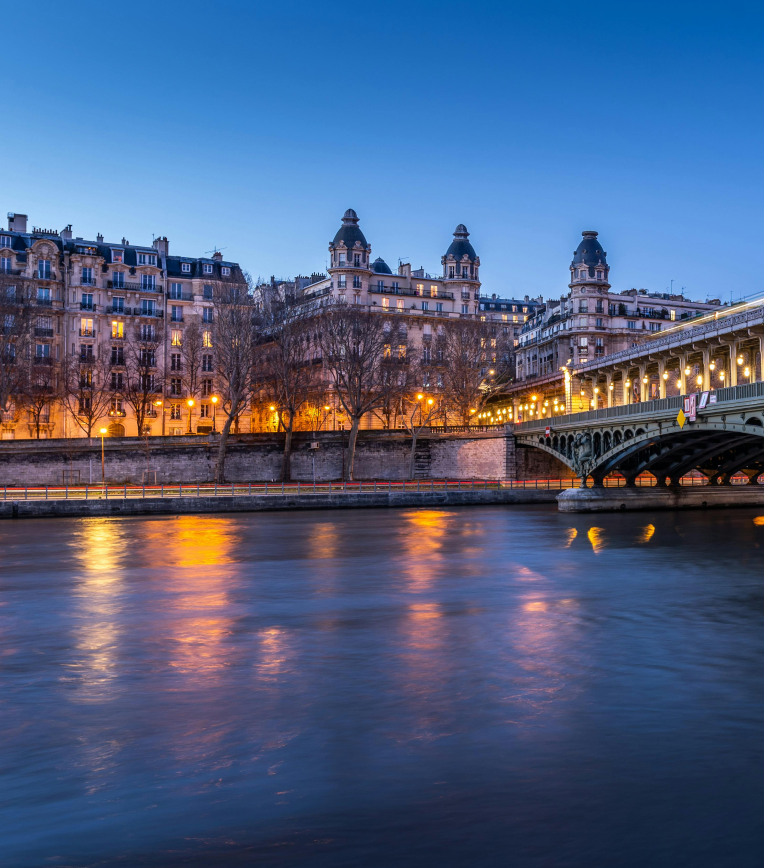 Evening view of historic Paris buildings along the River Seine with illuminated bridge and reflections on the water.