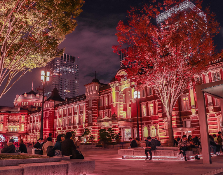 Tokyo Station Marunouchi building illuminated at night with people relaxing in the plaza and city towers in the background.