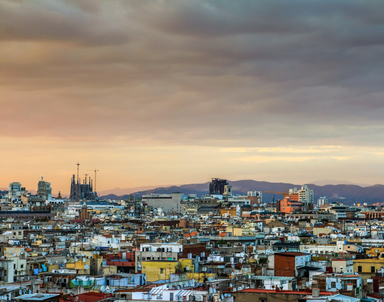 Barcelona skyline at sunset with Sagrada Familia rising above the cityscape, colorful rooftops in the foreground and mountains in the background.