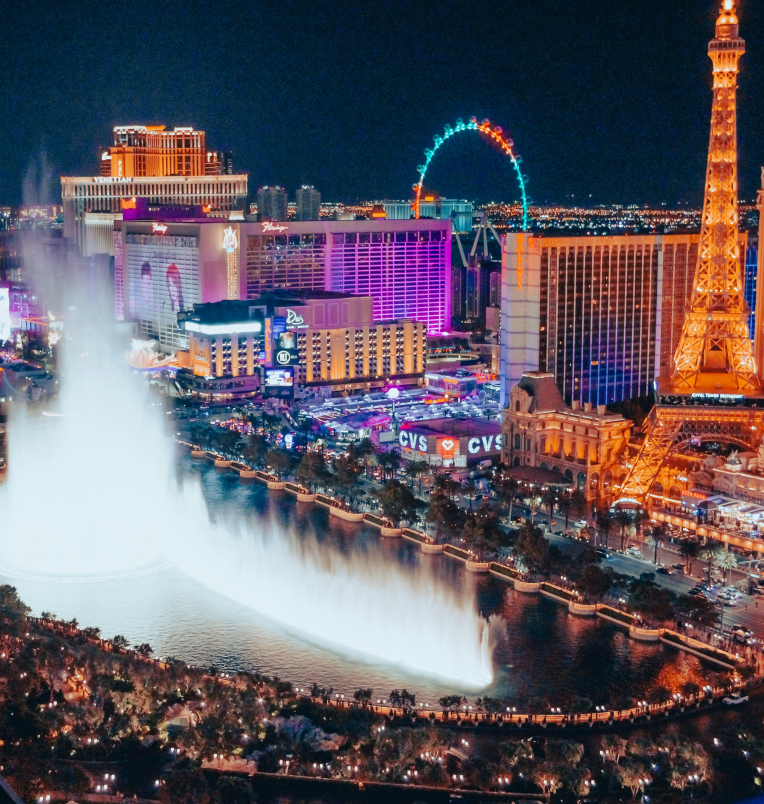 Bellagio Fountains show at night on the Las Vegas Strip with illuminated hotels, Paris Las Vegas Eiffel Tower replica, and the High Roller observation wheel in the background.