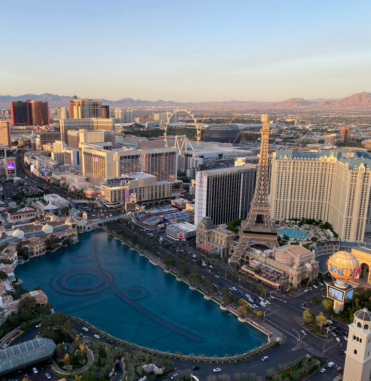 Aerial view of the Las Vegas Strip featuring the Bellagio fountains, Paris Las Vegas Eiffel Tower replica, and surrounding hotels against the Nevada desert backdrop at sunset.