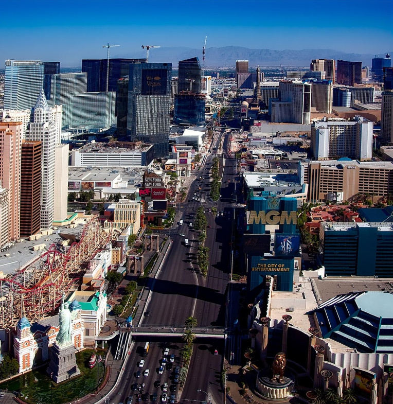 Aerial view of the Las Vegas Strip featuring MGM Grand, New York-New York Hotel roller coaster, and iconic casino resorts along Las Vegas Boulevard under a clear blue sky.