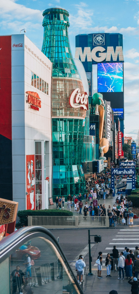Crowds walking along the Las Vegas Strip near the MGM Grand and Coca-Cola Store during the day with retail shops and casino signage visible along Las Vegas Boulevard.