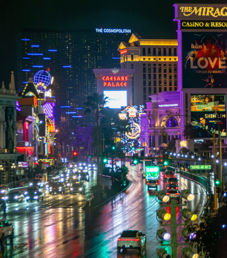 Las Vegas Strip at night with neon lights, Caesars Palace and The Mirage signage, and traffic reflecting on wet Las Vegas Boulevard.