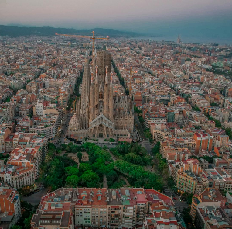 Aerial view of Sagrada Familia in Barcelona surrounded by the Eixample district, showcasing Antoni Gaudí’s iconic basilica rising above the city skyline at dusk.