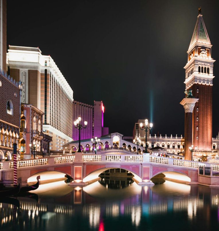 The Venetian Resort in Las Vegas at night featuring the Grand Canal bridge, illuminated campanile tower, and reflections on the water along the Strip.