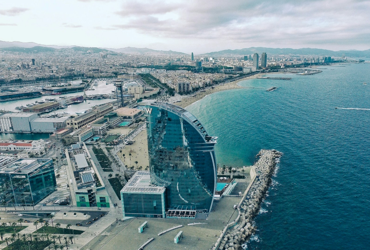 Aerial view of W Barcelona Hotel at Barceloneta Beach, featuring the sail-shaped glass tower along the Mediterranean coastline and Barcelona city skyline.