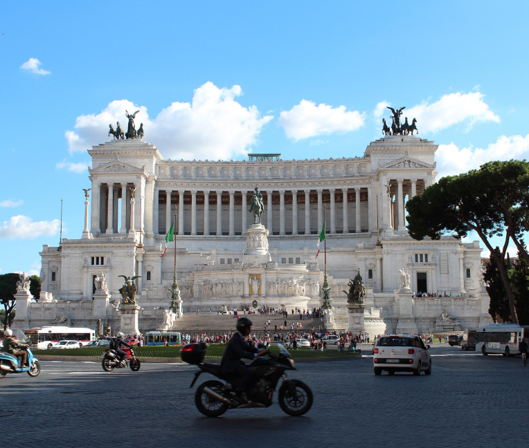 Altare della Patria (Vittoriano) monument in Rome at Piazza Venezia with Italian flags, statues, and traffic in foreground under blue sky.