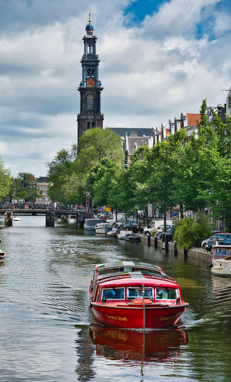 Where to Stay in Amsterdam. Red canal boat cruising through Amsterdam canal with Westerkerk tower in the background, historic houses and trees lining the water.