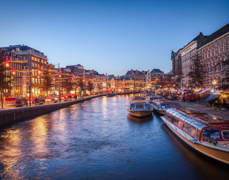 Evening view of an Amsterdam canal with illuminated historic buildings, canal boats and reflections on the water in the Netherlands.