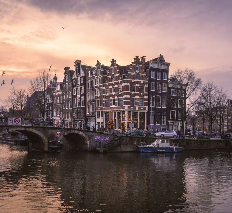 Historic canal houses in Amsterdam at sunset with stone bridge, boats and reflections on the water in the Netherlands.