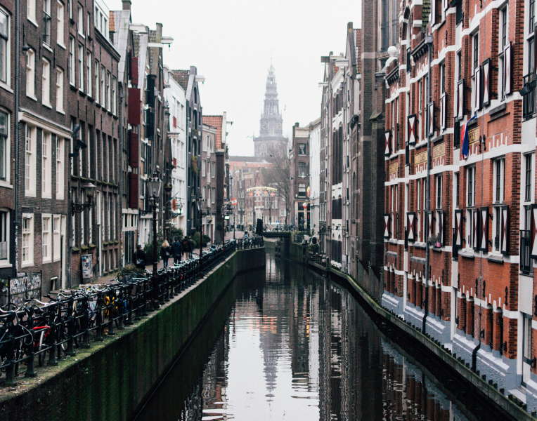 Narrow Amsterdam canal lined with traditional Dutch houses and bicycles, with Westerkerk tower visible in the background, Netherlands.