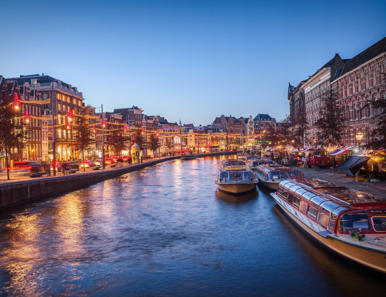 Amsterdam canal at night with illuminated historic buildings, boats docked along the water and city lights reflecting on the canal.