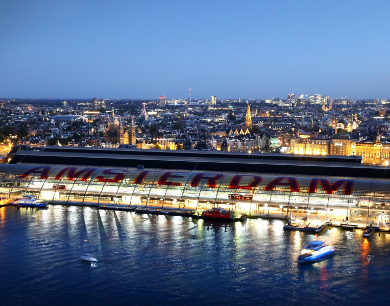 Amsterdam Central Station illuminated at night with city skyline and boats on the IJ waterfront in the Netherlands.