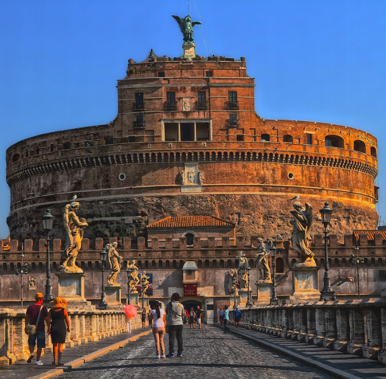 Castel Sant’Angelo in Rome at sunset, viewed from Ponte Sant’Angelo with statues, cobblestone bridge and tourists walking toward the historic fortress.