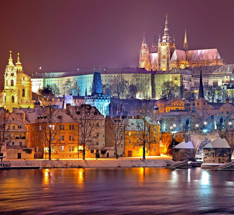 Prague Castle illuminated at night above the Vltava River with snow-covered rooftops and colourful Old Town buildings in winter, Czech Republic.