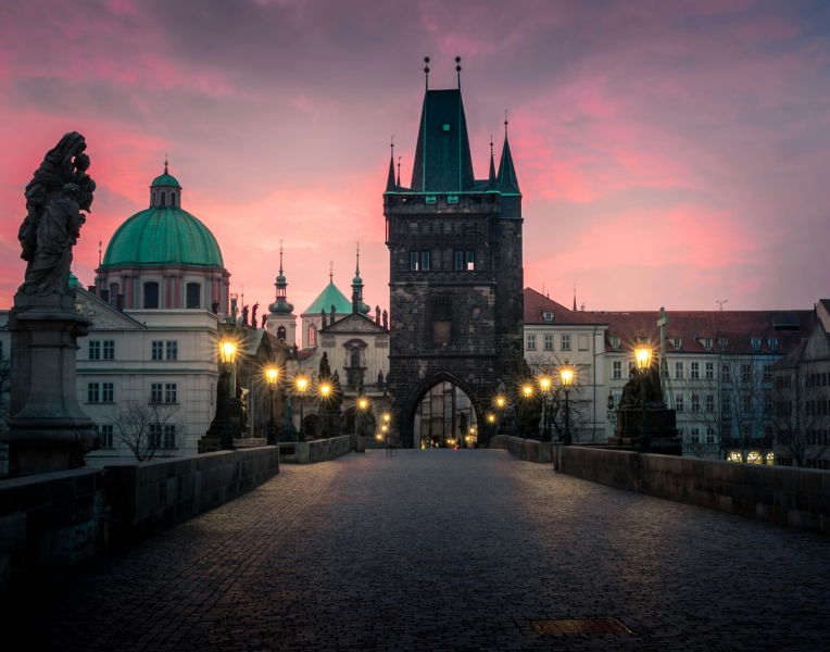 Charles Bridge in Prague at sunrise with the Old Town Bridge Tower and historic church domes under a pink sky, Czech Republic.