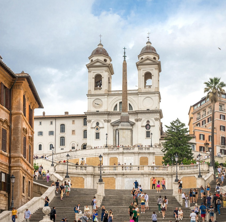 Spanish Steps in Rome at Piazza di Spagna with Trinità dei Monti church and obelisk, tourists walking on the staircase under cloudy sky.