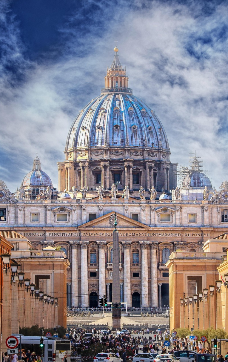 Where to Stay in Rome. St Peter’s Basilica dome in Vatican City, Rome, viewed from St Peter’s Square with historic columns and crowd under blue sky.