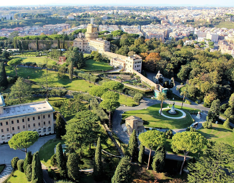 Aerial view of the Vatican Gardens in Vatican City with manicured lawns, trees, historic buildings and Rome skyline in the background.